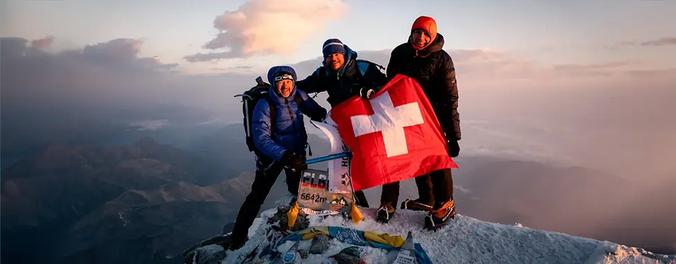Drei Bergsteiger von Air-Davos mit Schweizer Fahne auf dem Gipfel des Elbrus (5642 m) bei Sonnenaufgang